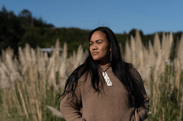A woman with long dark hair wearing a brown sweater and a carved necklace stands outdoors in front of tall grasses, looking thoughtfully into the distance under a clear blue sky.